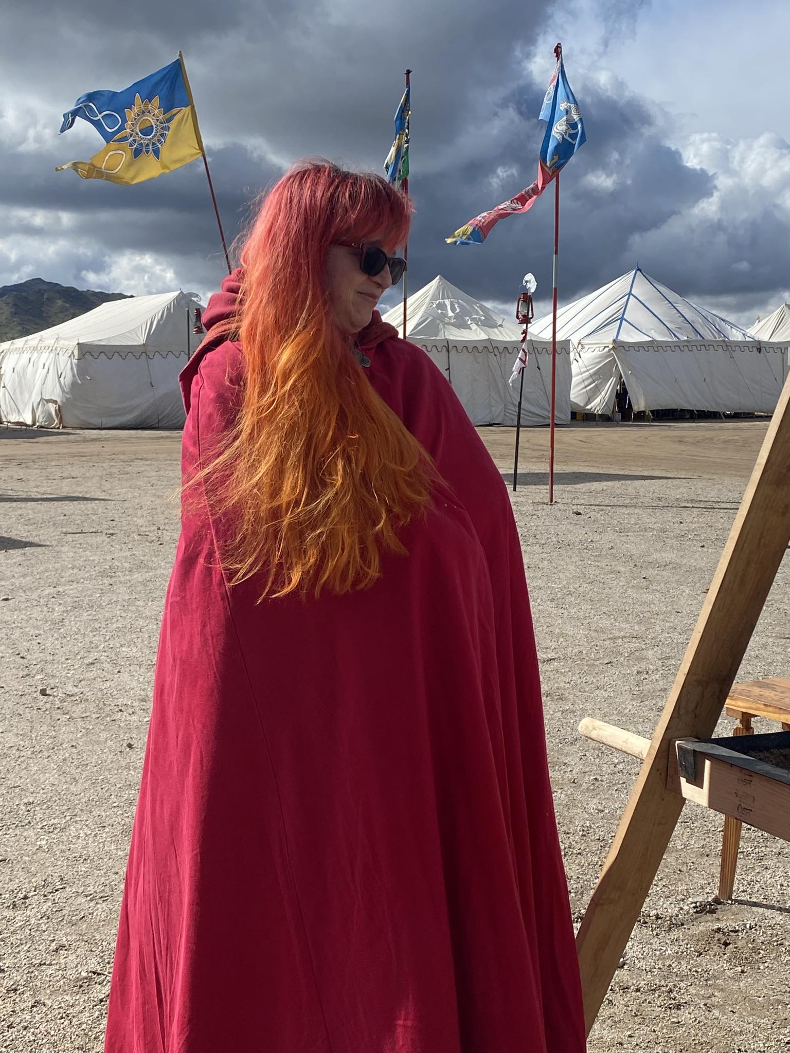 A lady of the Kingdom posed with her long auburn hair draped over her woolen red cloak. Windy clouds and banners can be seen in the distance.