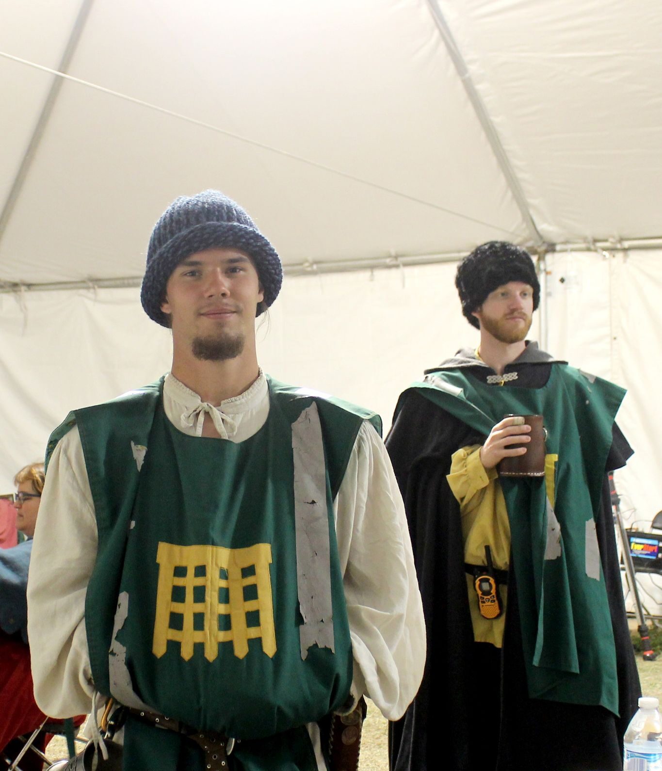 Two younger male presenting populace members wearing green tabbards with The Watch heraldry embroidered and displayed in gold above their attire. Both wearing cold weather tunics and caps.